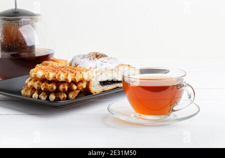 Tazza di tè, piattino con pasticceria e teiera rotonda trasparente su tavolo di legno bianco. Foto Stock