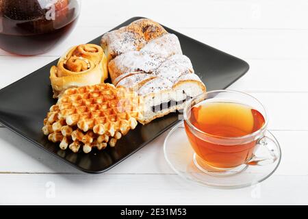 Tazza di tè, piattino con pasticceria e teiera rotonda trasparente su tavolo di legno bianco. Foto Stock