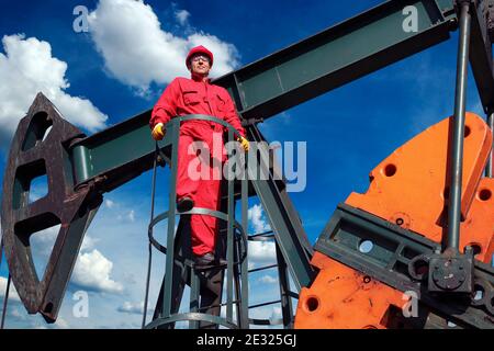 Operaio in tute rosse e indumenti protettivi personali in piedi al Pump Jack Pump Unit. Operaio di petrolio in un campo petrolifero contro il cielo blu. Foto Stock