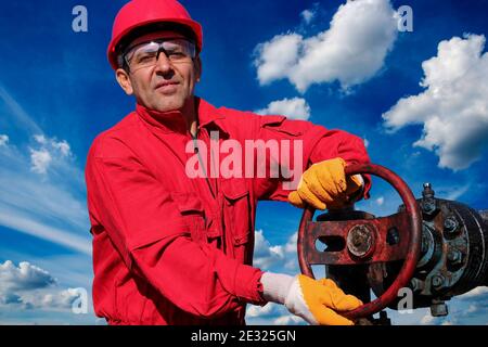 Ritratto di un lavoratore contro il cielo blu con le nuvole bianche al pozzo di petrolio di Jack di pompa. Operaio di petrolio in tute rosse e indumenti protettivi personali. Foto Stock