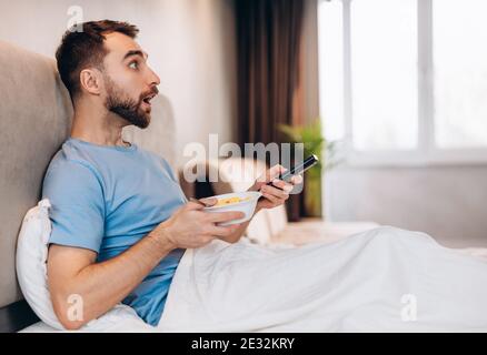 Attraente giovane uomo con barba che mangia la colazione a letto e guarda sorpreso scioccato mentre guarda le notizie. Foto Stock