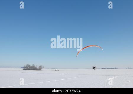 Parapendio motorizzato arancione che vola in un campo innevato in inverno con orizzonte e cielo blu Foto Stock