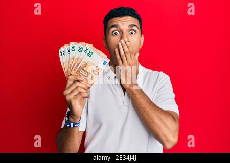 Giovane arabo con 50 banconote in euro che coprono la bocca con la mano, scioccato e impaurito per gli errori Foto Stock