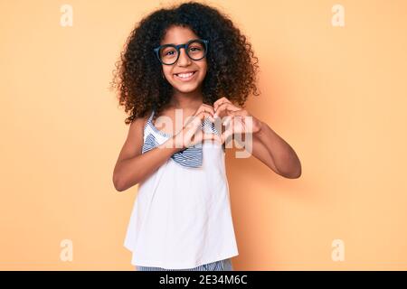 Bambino afroamericano con capelli ricci che indossa abiti casual e occhiali sorridenti in amore facendo il simbolo del cuore forma con le mani. Concetto romantico. Foto Stock