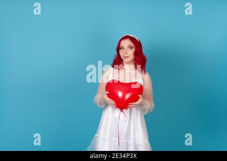 una giovane donna sorridente in un vestito bianco con i capelli rossi trattengono un pallone volante a forma di cuore isolato su un sfondo blu con entrambe le mani Foto Stock