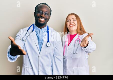 Giovane coppia interracial che indossa uniforme medico e stetoscopio sorridente amichevole offrendo handshake come saluto e accogliente. Business di successo. Foto Stock