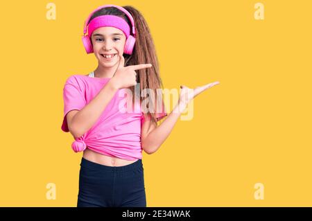 Carina bambina ispanica che indossa abiti da palestra e utilizza le cuffie stupito e sorridente alla fotocamera mentre si presenta con la mano e puntando con il dito Foto Stock