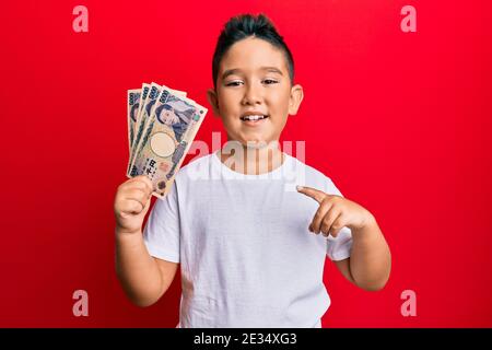 Ragazzino ispanico capretto che tiene 5000 biglietti giapponesi dello yen sorridenti buon puntamento con mano e dito Foto Stock