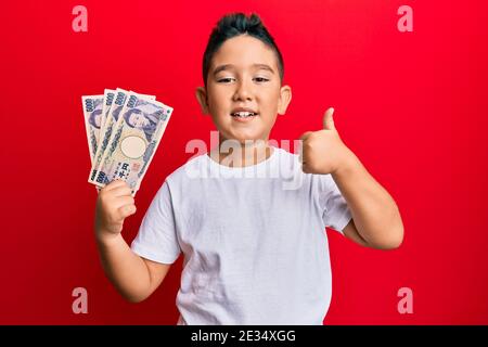 Bambino ispanico ragazzo piccolo che tiene 5000 yen giapponesi banconote sorridente felice e positivo, pollice su facendo eccellente e segno di approvazione Foto Stock