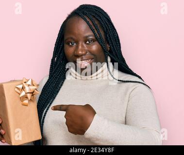 Giovane donna nera con trecce che tengono il regalo sorridente che punta felice con mano e dito Foto Stock