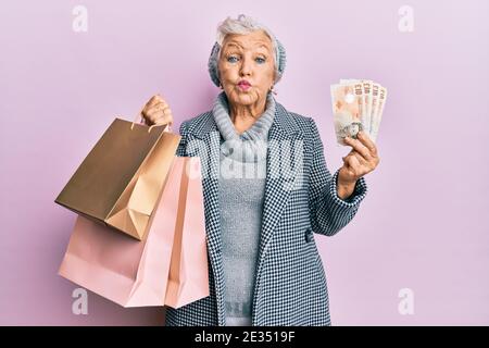 Donna anziana con capelli grigi che tiene borse per la spesa e banconote inglesi che soffia guance con viso divertente. Bocca gonfia con aria, cattura aria. Foto Stock