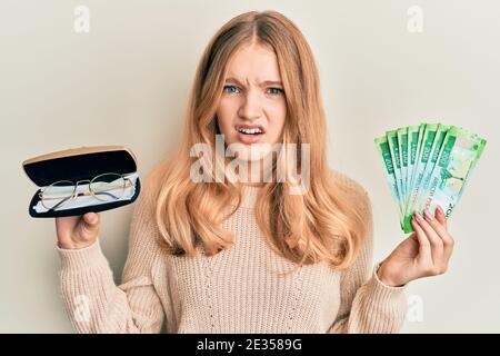 Bella giovane caucasica ragazza che tiene bicchieri e le banconote rubate russe in faccia d'urto, guardando scettico e sarcastico, sorpreso con la bocca aperta Foto Stock