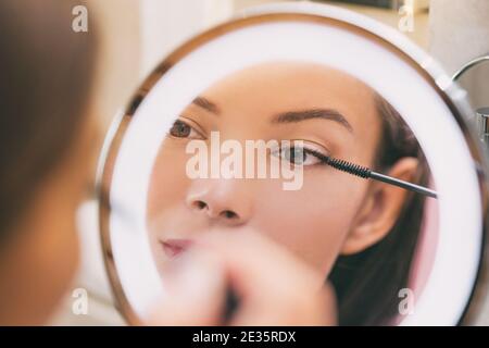 Donna che applica il prodotto di bellezza che mette il mascara nell'anello illuminato specchio rotondo di trucco alla procedura domestica del mattino del bagno. Bella signora asiatica Foto Stock