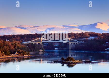 Vista panoramica sulle montagne innevate di Snowdonia attraverso lo stretto di Menai con il ponte sospeso di Menai in inverno. Menai Bridge Isola di Anglesey Wales Gran Bretagna Foto Stock