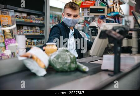 Giovane addetto alle vendite che indossa la maschera di protezione seduta presso il registratore di cassa nel supermercato e gli acquirenti di servizio. Cassa maschile scansione prodotti alimentari al momento del pagamento. Foto Stock