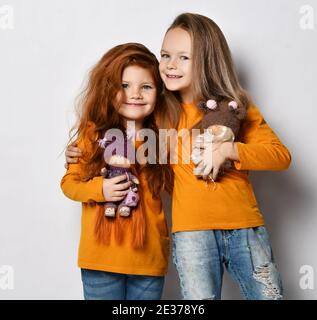 Ritratto di due ragazze felici di bambini migliori amici in simile i vestiti si levano in piedi insieme con le loro bambole favorite che abbracciano Foto Stock