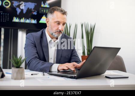 Bell'uomo d'affari fidato e convinto in abiti formali, seduto al tavolo in un moderno ufficio leggero, digitando sul laptop, lavorando Foto Stock