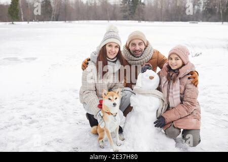 Ritratto a lunghezza intera di felice famiglia che costruisce pupazzo di neve insieme all'aperto e guardando la macchina fotografica mentre godendo la vacanza invernale, copy space Foto Stock