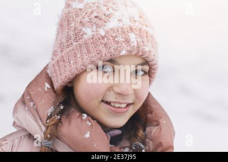 Primo piano ritratto di ragazza carina in inverno che indossa cappello rosa e giacca mentre si guarda la fotocamera coperta di neve, copia spazio Foto Stock