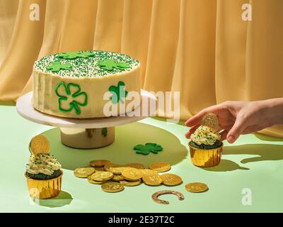 Concetto di cibo per il giorno di San Patrizio. Moderna vita morta con cibo dolce per la festa di San Patrizio con la mano femminile. Sciamrocks decorato con torte, cupcake di velluto verde, monete d'oro al cioccolato e ferro di cavallo Foto Stock