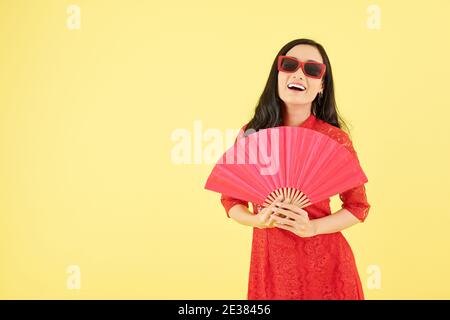 Bella giovane donna ridente in occhiali da sole e vestito rosso in posa con ventola della carta Foto Stock