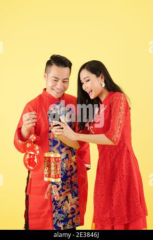 Entusiasta giovane asiatica che mostra foto sullo schermo dello smartphone al ragazzo quando stanno tenendo le decorazioni con i migliori auguri iscrizione Foto Stock
