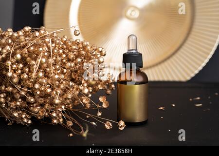 Bottiglia di olio essenziale o siero facciale con pipetta e fiori d'oro su sfondo nero. Vista frontale Foto Stock