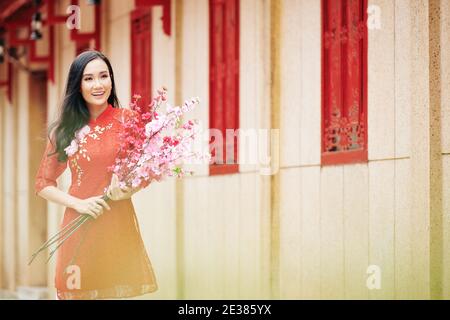 Attraente giovane donna asiatica sorridente in abito rosso in piedi all'aperto con rami di pesca in fiore Foto Stock