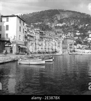 1950s, storico, una vista del grazioso villaggio costiero, Villefranche-sur-Mer nel sud della Francia in questa epoca. Una pittoresca città vecchia situata nella regione delle Alpi-Costa Azzurra sulla Costa Azzurra, Villefranche è considerata una delle baie più belle del mondo e circondata dal Cap di Nizza e Cap Ferrat. Nella foto si vede il bar/ristorante, il porto Nautico e recintato. Foto Stock