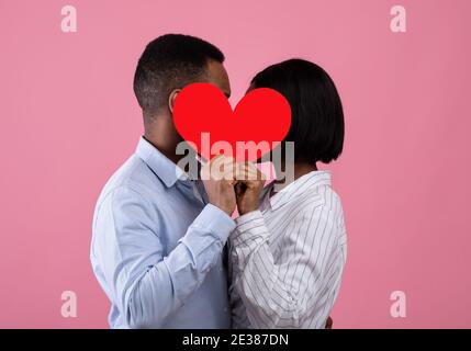 Vista laterale di appassionati amanti neri che baciano dietro il cuore di carta rossa, esprimendo il loro amore su sfondo rosa studio Foto Stock