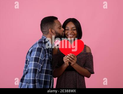 Festa di San Valentino. Bel ragazzo nero baciare la sua bella ragazza con cuore rosso su sfondo rosa Foto Stock