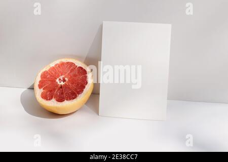 Moderna stationeria estiva ancora scena di vita. Closeup di pompelmo tagliato su sfondo bianco della tavola alla luce del sole. Carta bianca, mockup invito Foto Stock
