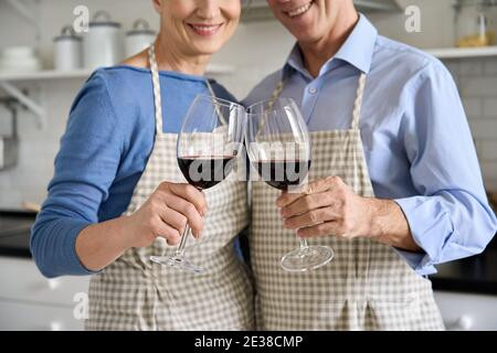 Felice coppia senior che tiene bicchieri bere vino in piedi in cucina, primo piano. Foto Stock