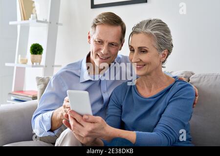 Coppia di mezza età che guarda lo smartphone utilizzando le app mobili siedono sul divano a casa. Foto Stock