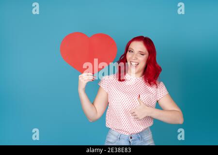 ritratto di una giovane maliziosa e giocosa con capelli rossi che reggono un cuore di carta rosso, che si avvolge e dà un pollice in su, isolata su uno sfondo blu Foto Stock