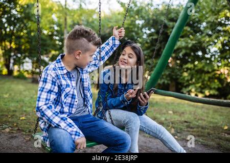 Felici i bambini sorridenti che guardano video divertenti sullo smartphone mentre si siedono in swing. Messa a fuoco selettiva. Foto Stock