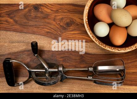 Uno sfondo a tema alimentare con una ciotola di uova multicolore su un tagliere di legno con un miscelatore manuale. L'accoppiatore sinistro è aperto per il testo. Foto Stock