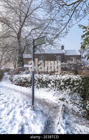 Un cartello per un ponte pubblico a Baillon, Yorkshire, Inghilterra. L'immagine è una scena invernale con un cottage innevato e alberi sullo sfondo. Foto Stock