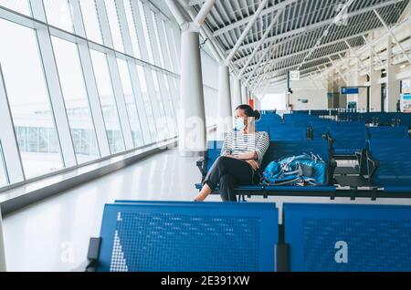 Solitario viaggiatore femminile con zaino seduto nella sala di trasferimento passeggeri vuota dell'aeroporto in maschera protettiva e guardando verso l'esterno grandi finestre. Foto Stock