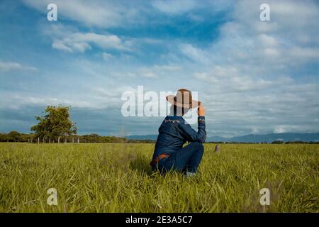 Un fuoco poco profondo di una femmina vestita da cowboy seduto a terra in un campo sotto un blu cielo nuvoloso Foto Stock