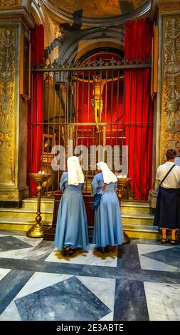 Due monache visitano una piccola cappella a Roma, in Italia, sotto una statua di Gesù Cristo sulla Croce, illuminata per creare un'ombra sul muro dietro Foto Stock