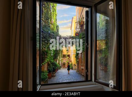 Vista attraverso una finestra aperta mentre una giovane donna cammina attraverso un vicolo colorato coperto di edera nel quartiere Trastevere di Roma, Italia Foto Stock