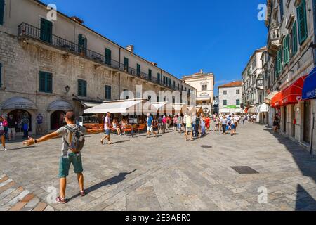 Un uomo posa per una foto con le braccia allungate dentro La Piazza delle armi nella città medievale di Kotor Montenegro come turisti a piedi da caffè e negozi Foto Stock