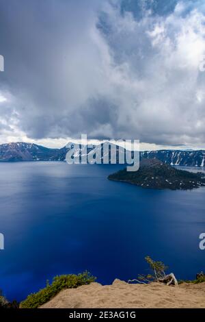 Splendido scenario al lago Crater. Isola di Wizard nel mezzo. Catena montuosa innevata intorno al bordo. Cielo nuvoloso e blu. Lago profondo, blu scuro. Foto Stock