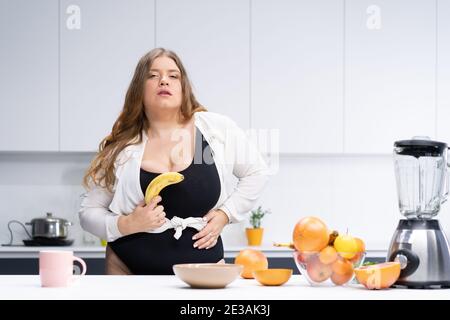 Concetto di dieta e nutrizione. Giovane donna dal corpo curvo felice con lunghi capelli biondi usando il frullatore nella cucina moderna, mescolando la frutta fresca per sano Foto Stock