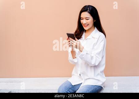 Donna asiatica testando su smartphone mentre si siede su una panca di cemento circondata da pentole di piante verdi con una parete di sabbia deserta colorata sullo sfondo e foglie verdi in primo piano. Foto Stock