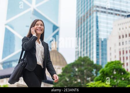 Donna d'affari asiatica che parla sul telefono cellulare che cammina nella strada di Hong Kong per l'ufficio, grattacieli paesaggio urbano sfondo. Giovane donna su smartphone Foto Stock