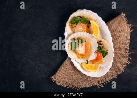 Capesante al forno con caviale e salsa cremosa all'aglio in piatto bianco. Capesante con limone su fondo nero con tovagliolo. Vista dall'alto. Spazio di copia. Disposizione piatta Foto Stock