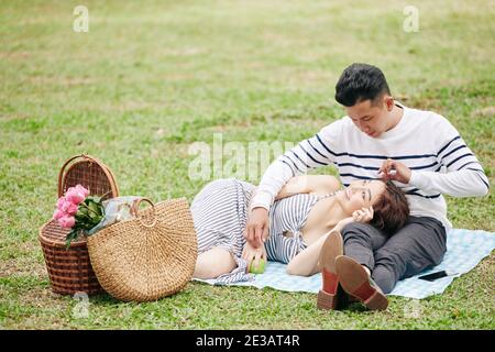Giovane uomo vietnamita innamorato guardando la testa di appoggio della ragazza sui suoi giri quando stanno avendo picnic nel parco Foto Stock