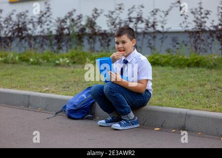 scolaro in una camicia bianca con cravatta blu, tiene una scatola blu per il pranzo e mangia un pezzo di mela Foto Stock